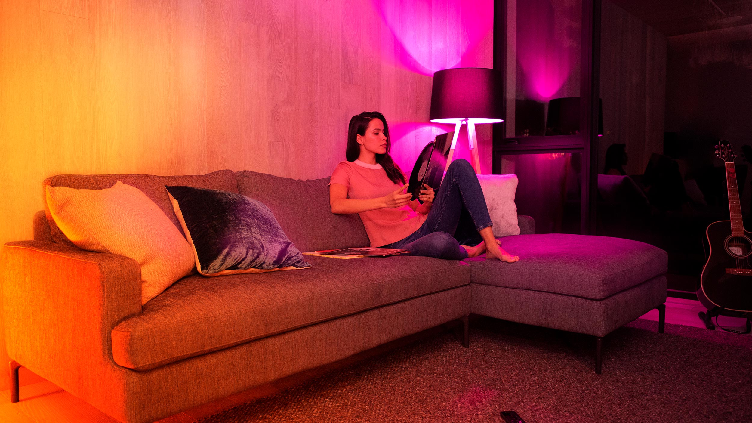 A woman relaxing on a modern sectional sofa, illuminated by vibrant colored lighting, while holding a vinyl record, creating a cozy and artistic atmosphere.